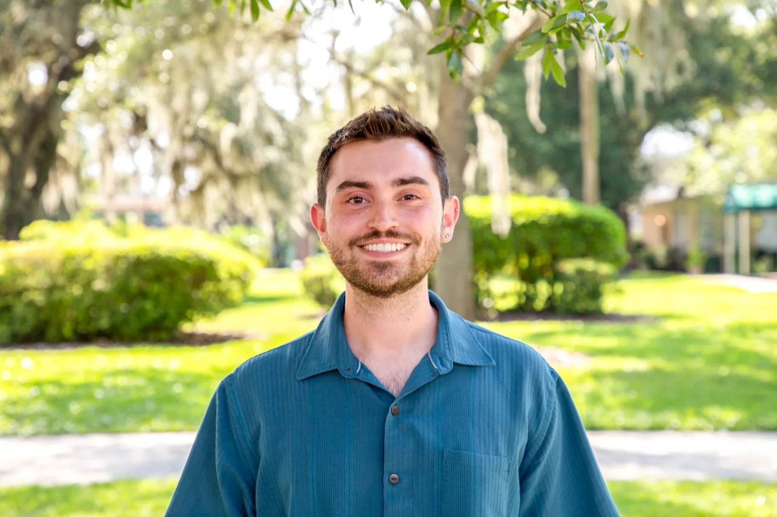 Wesley Chandler smiling outdoors in a blue shirt against a sunlit green background